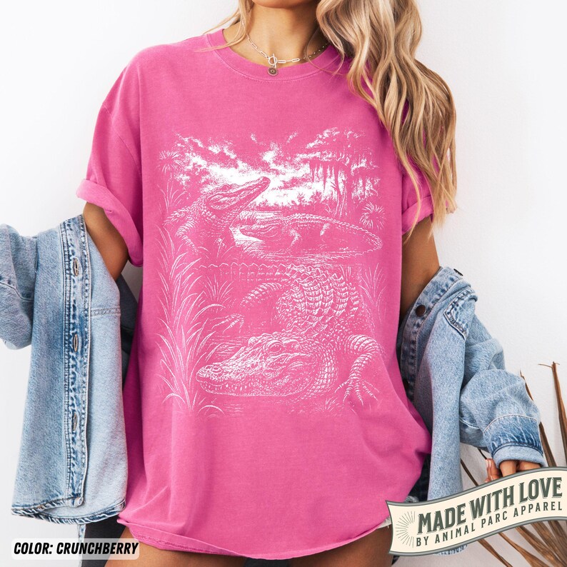 May include: A pink t-shirt with a white graphic of alligators in a swamp scene. The shirt is a solid color, with the text "MADE WITH LOVE BY ANIMAL PARC APPAREL" in a banner on the bottom right. The color is "CRUNCHBERRY".