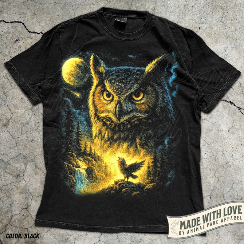 May include: Black t-shirt with a detailed illustration of an owl, a full moon, and a small bird. The design incorporates shades of blue, yellow, and black. The text "MADE WITH LOVE BY ANIMAL PARC APPAREL" is visible. The shirt is a standard fit.
