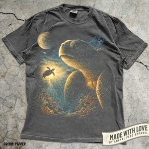 May include: Pepper-colored t-shirt featuring a detailed illustration of a sea turtle with a smaller turtle swimming towards a bright light. The design includes a moon and coral. The shirt has the text "MADE WITH LOVE BY ANIMAL PARC APPAREL".