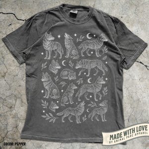 Pode incluir: Camiseta cinza-pimenta com uma ilustração branca de lobos, estrelas e luas crescentes. O design inclui várias poses de lobos e elementos botânicos. O texto "MADE WITH LOVE BY ANIMAL PARC APPAREL" está impresso em uma faixa.