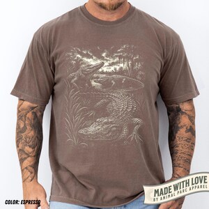 May include: Espresso-colored t-shirt featuring a detailed illustration of alligators in a swamp setting. The design includes multiple alligators, tall grass, and a cloudy sky. The shirt has short sleeves and a relaxed fit. The text "MADE WITH LOVE BY ANIMAL PARC APPAREL" is printed on the bottom right.