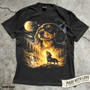 May include: Black t-shirt featuring a detailed illustration of a dachshund dog under a full moon, with a waterfall and forest scene. The dog is depicted in a realistic style with brown and black fur. The shirt includes the text "MADE WITH LOVE BY ANIMAL PARC APPAREL".