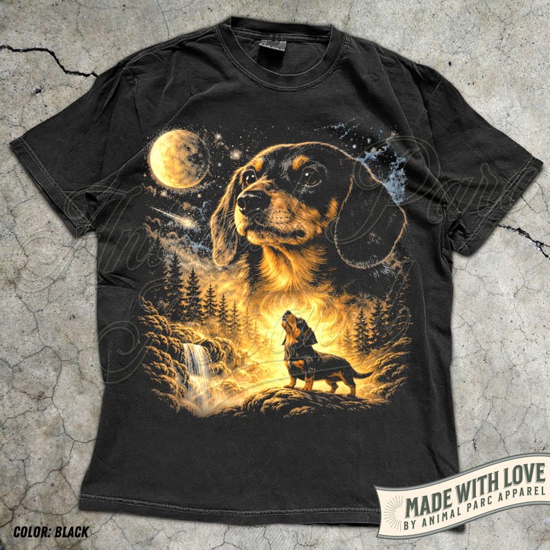 May include: Black t-shirt featuring a detailed illustration of a dachshund dog under a full moon, with a waterfall and forest scene. The dog is depicted in a realistic style with brown and black fur. The shirt includes the text "MADE WITH LOVE BY ANIMAL PARC APPAREL".