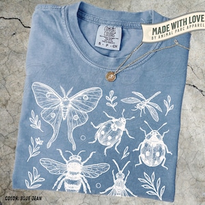 May include: A blue jean coloured t-shirt featuring white insect illustrations, such as a moth, dragonfly, and bee. The shirt has a crew neck and the text "COMFORT COLORS" on the tag. A gold necklace with a pendant is on the shirt. The text "MADE WITH LOVE" is also visible.