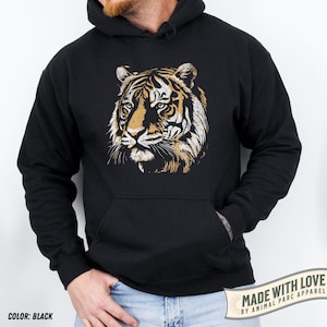 May include: Black hoodie featuring a detailed tiger face graphic in shades of brown, tan, and white. The hoodie has a kangaroo pocket and a drawstring hood. The text "COLOR: BLACK" is at the bottom left.