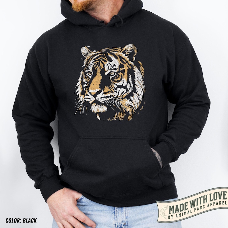 May include: Black hoodie featuring a detailed tiger face graphic in shades of brown, tan, and white. The hoodie has a kangaroo pocket and a drawstring hood. The text "COLOR: BLACK" is at the bottom left.