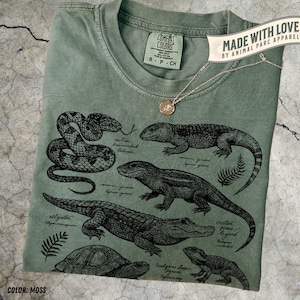 May include: A moss green t-shirt featuring a black illustration of reptiles and amphibians, such as a snake, alligator, and lizard. The shirt has a crew neck and a small tag that says "Comfort Colors". A gold necklace with a pendant is draped across the shirt. The text "Made with Love by Animal Parc Apparel" is also visible.