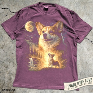 May include: Berry-colored t-shirt featuring a detailed illustration of a Corgi dog with a celestial background, including a moon, stars, and a waterfall. The design also includes a smaller Corgi and trees. The text "MADE WITH LOVE BY ANIMAL PARC APPAREL" is visible.