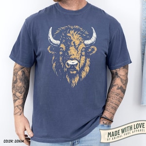May include: Denim blue t-shirt featuring a detailed illustration of a bison's head in shades of gold and white. The shirt includes the text "MADE WITH LOVE BY ANIMAL PARC APPAREL" and "COLOR: DENIM".