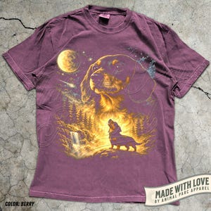 May include: Berry-colored t-shirt featuring a detailed illustration of a dachshund dog under a night sky with a full moon, waterfall, and forest. The design includes the text "MADE WITH LOVE BY ANIMAL PARC APPAREL" and "COLOR: BERRY".