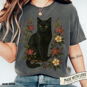 Könnte beinhalten: Graues T-Shirt mit einem schwarzen Katzendesign, umgeben von roten und gelben Blumen und grünen Blättern. Das Shirt hat den Text "MADE WITH LOVE BY ANIMAL PARC APPAREL" und "COLOR: PEPPER".