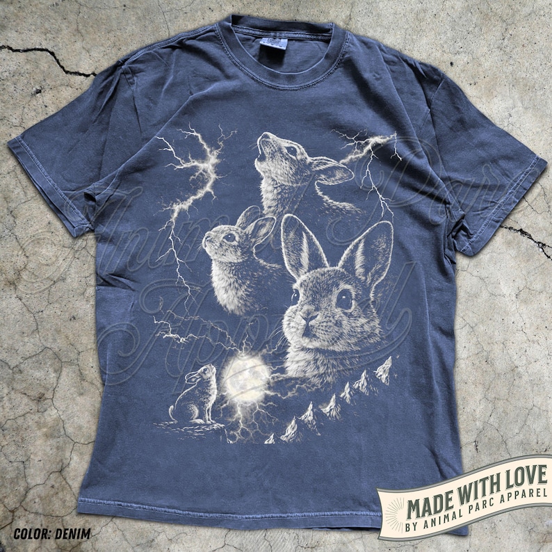 Pode incluir: Camiseta azul denim com uma ilustra&ccedil;&atilde;o de coelhos, raios e lua cheia. O design inclui o texto "MADE WITH LOVE BY ANIMAL PARC APPAREL" em uma faixa. A camiseta &eacute; de gola redonda e manga curta.