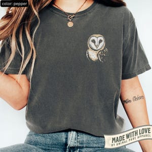 Può includere: T-shirt color pepe con grafica dettagliata di un gufo sul petto. La maglietta ha una vestibilità ampia e maniche corte. Il testo "MADE WITH LOVE BY ANIMAL PARC APPAREL" è stampato su uno striscione in basso.