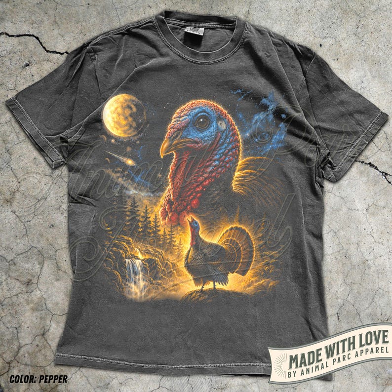 Puede incluir: Una camiseta de manga corta gris carb&oacute;n con un estampado gr&aacute;fico de una cabeza de pavo grande y un pavo m&aacute;s peque&ntilde;o en un paisaje forestal con una cascada, una luna amarilla brillante y una estrella fugaz sobre un fondo c&oacute;smico oscuro.