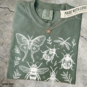 以下が含まれることがあります： モスグリーンのTシャツに、白い昆虫と植物のイラスト。デザインには蛾、トンボ、蜂、その他の昆虫が含まれています。ペンダント付きのゴールドネックレスがTシャツの上にあります。「Comfort Colors」と「Made with Love」と書かれたタグが付いています。