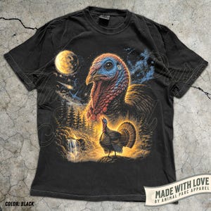 Puede incluir: Una camiseta negra con un estampado gr&aacute;fico que muestra la cabeza de un pavo grande en la parte superior y un pavo m&aacute;s peque&ntilde;o de pie sobre una roca cerca de una cascada y pinos debajo. Una luna amarilla y una estrella fugaz aparecen en el cielo.