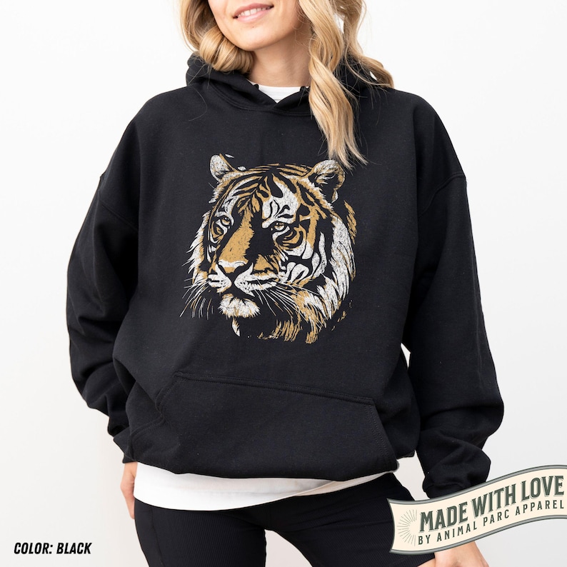 May include: Black hooded sweatshirt featuring a detailed tiger face graphic in shades of gold, white, and brown. The sweatshirt has a kangaroo pocket and a drawstring hood. The text "COLOR: BLACK" is at the bottom left.
