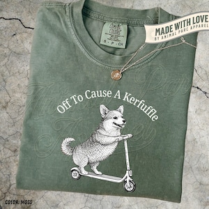 May include: Moss green t-shirt with a graphic of a corgi on a scooter and the text "Off To Cause A Kerfuffle." The shirt has a crew neck and short sleeves. A gold-colored necklace with a pendant is draped across the shirt.