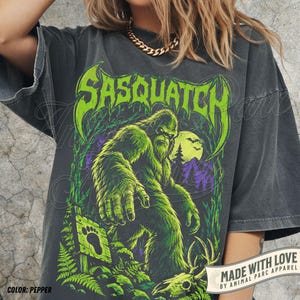 May include: A charcoal gray t-shirt featuring a large graphic print of a green Sasquatch in a forest setting. The text "SASQUATCH" appears in a stylized green font above the creature. The design includes a wooden sign with a footprint icon.