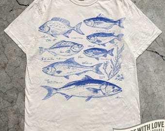 Puede incluir: Camiseta de color marfil con ilustraciones azules de varias especies de peces y sus nombres. La camiseta incluye el texto "MADE WITH LOVE BY ANIMAL PARC APPAREL" y el color se indica como "IVORY".