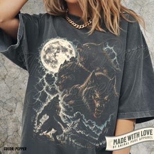 May include: A charcoal grey oversized t-shirt featuring a graphic print of three snarling wolves, a full moon, lightning bolts, and a mountain landscape. The shirt has short sleeves and a crew neckline. The text "MADE WITH LOVE BY ANIMAL PARC APPAREL" is visible.