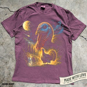 Puede incluir: Una camiseta de manga corta color baya con un estampado gr&aacute;fico de una cabeza de pavo grande y un pavo m&aacute;s peque&ntilde;o de pie en un paisaje forestal con una cascada, una luna amarilla brillante y una estrella fugaz en un cielo nocturno estrellado.