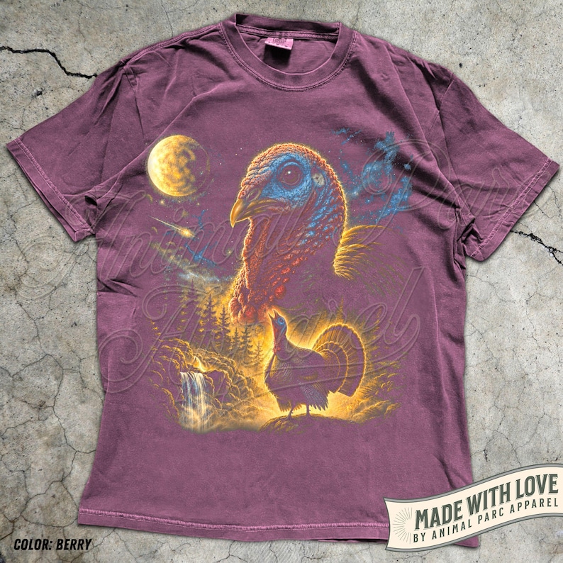 Puede incluir: Una camiseta de manga corta color baya con un estampado gr&aacute;fico de una cabeza de pavo grande y un pavo m&aacute;s peque&ntilde;o de pie en un paisaje forestal con una cascada, una luna amarilla brillante y una estrella fugaz en un cielo nocturno estrellado.