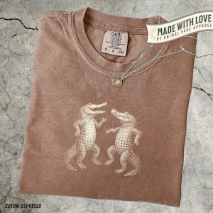 May include: Espresso-coloured t-shirt with a graphic of two alligators. The shirt has a crew neck and a tag that reads "COMFORT COLORS". A silver necklace with a pendant rests on the shirt. The text "MADE WITH LOVE BY ANIMAL PARC APPAREL" is also visible.