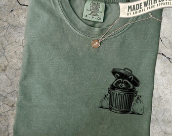 May include: Moss green t-shirt with a raccoon in a trash can graphic. The shirt has the words "Comfort Colors" and "Made with Love by Animal Parc Apparel" printed on it. A gold necklace with a small pendant is on the shirt.