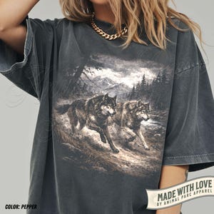 K&ouml;nnte beinhalten: Pfefferfarbenes T-Shirt mit einer detaillierten Grafik von zwei W&ouml;lfen, die durch eine Waldlandschaft laufen. Das Design umfasst Berge und B&auml;ume im Hintergrund. Das Shirt hat kurze &Auml;rmel und eine lockere Passform. Der Text "MADE WITH LOVE BY ANIMAL PARC APPAREL" ist sichtbar.