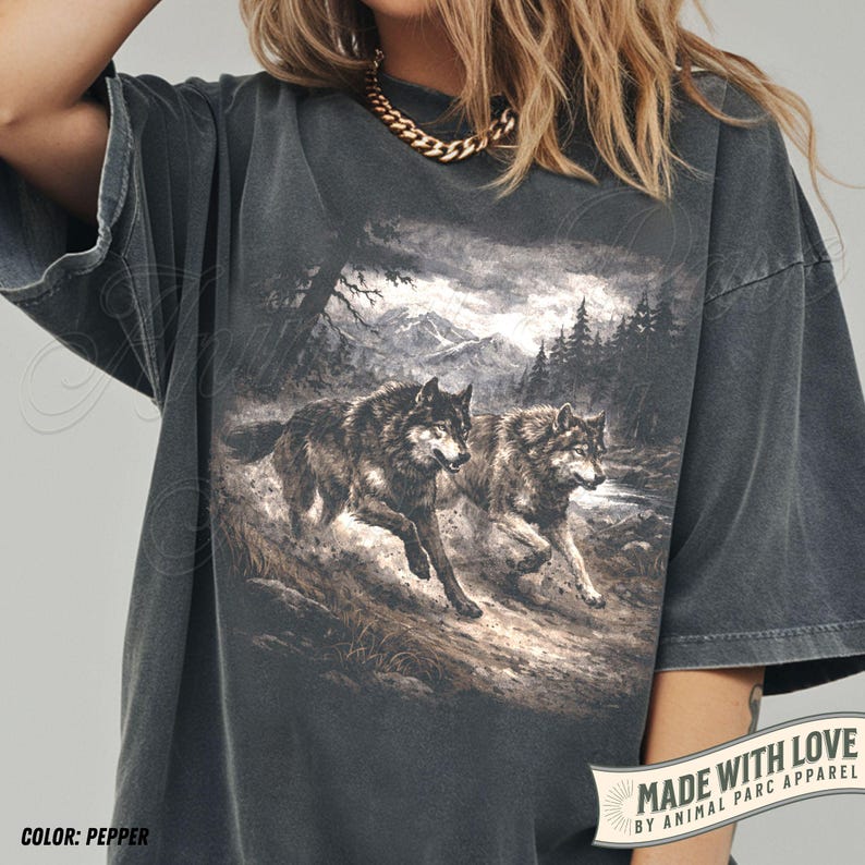 K&ouml;nnte beinhalten: Pfefferfarbenes T-Shirt mit einer detaillierten Grafik von zwei W&ouml;lfen, die durch eine Waldlandschaft laufen. Das Design umfasst Berge und B&auml;ume im Hintergrund. Das Shirt hat kurze &Auml;rmel und eine lockere Passform. Der Text "MADE WITH LOVE BY ANIMAL PARC APPAREL" ist sichtbar.