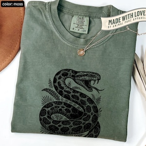 Peut inclure: T-shirt vert mousse Comfort Colors avec un motif de serpent noir détaillé. Le t-shirt a un col rond et des manches courtes. L'étiquette indique "Comfort Colors". Un collier doré avec un pendentif est posé sur le t-shirt. Le texte "MADE WITH LOVE BY ANIMAL PARC APPAREL" est visible.