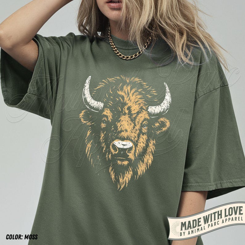 May include: Moss green t-shirt featuring a detailed illustration of a bison's head. The bison has tan fur and white horns. The shirt has short sleeves and a relaxed fit. The text "MADE WITH LOVE BY ANIMAL PARC APPAREL" is on the bottom right.