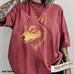 May include: Crimson-colored t-shirt featuring a detailed golden wolf head graphic. The shirt has a relaxed fit and short sleeves. The text "MADE WITH LOVE BY ANIMAL PARC APPAREL" is printed on a banner at the bottom. The color is crimson.