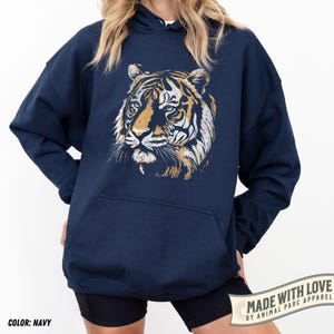 May include: Navy blue hoodie featuring a detailed tiger face graphic in shades of gold and white. The hoodie has a kangaroo pocket and a drawstring hood. The text "MADE WITH LOVE BY ANIMAL PARC APPAREL" is printed on a banner. The color is navy.