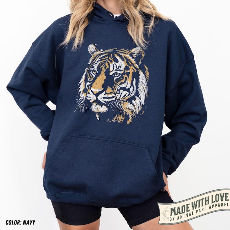 May include: Navy blue hoodie featuring a detailed tiger face graphic in shades of gold and white. The hoodie has a kangaroo pocket and a drawstring hood. The text "MADE WITH LOVE BY ANIMAL PARC APPAREL" is printed on a banner. The color is navy.