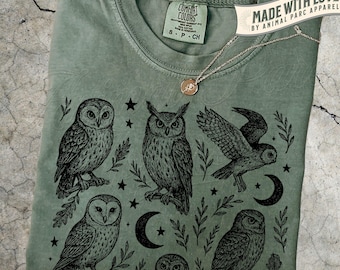 May include: Moss green t-shirt featuring a black illustration of several owls, stars, and crescent moons. The shirt has a crew neck and short sleeves. A silver necklace with a small pendant rests on the shirt. The tag reads "Comfort Colors".