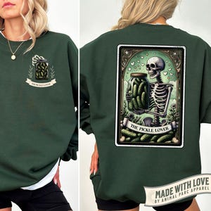 Pu&ograve; includere: Felpa verde scuro con colletto bianco. La parte anteriore presenta un barattolo di sottaceti con la scritta "THE PICKLE LOVER". Il retro mostra uno scheletro che tiene un barattolo di sottaceti, con lo stesso testo.