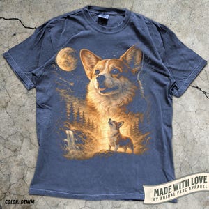 May include: Denim blue t-shirt featuring a detailed illustration of a Corgi dog with a forest and moon backdrop. The design includes a large Corgi portrait and a smaller one looking up. The text "MADE WITH LOVE" is visible on the bottom right.