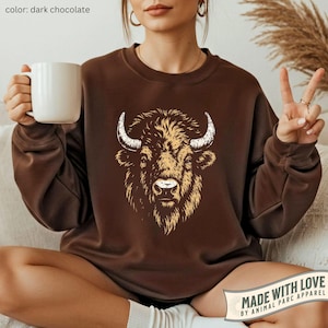May include: Dark chocolate-colored sweatshirt featuring a detailed bison graphic. The bison is depicted with tan fur and white horns. The sweatshirt is a crew neck style and has long sleeves. The text "MADE WITH LOVE BY ANIMAL PARC APPAREL" is visible.