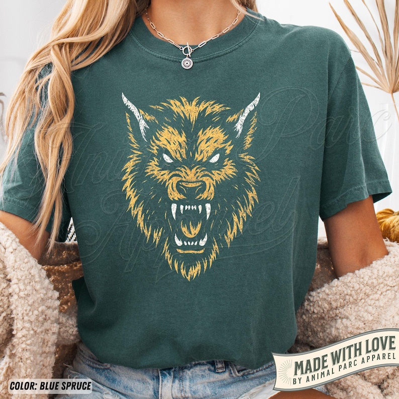 May include: A teal-coloured t-shirt with a yellow and white illustration of a wolf's head. The design shows a snarling wolf with sharp teeth. The text "MADE WITH LOVE BY ANIMAL PARC APPAREL" is printed on a banner at the bottom of the shirt.