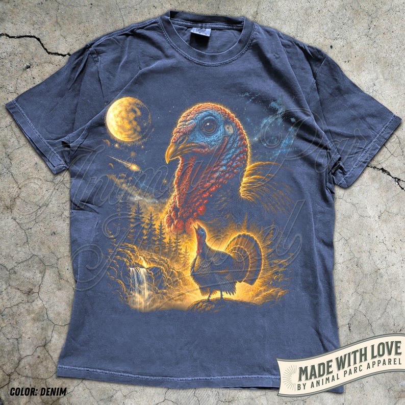Puede incluir: Una camiseta de mezclilla color carb&oacute;n con un estampado gr&aacute;fico de la cabeza de un pavo grande y un pavo m&aacute;s peque&ntilde;o de pie cerca de una cascada y un bosque. La escena incluye una luna amarilla brillante y una estrella fugaz en el cielo.