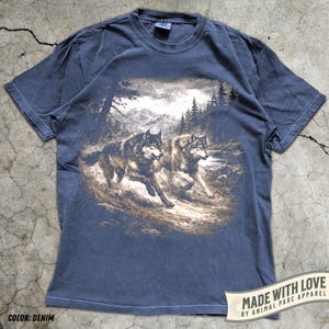 K&ouml;nnte beinhalten: Denimblaues T-Shirt mit einer Grafik von zwei W&ouml;lfen, die durch einen Wald laufen. Das Design umfasst B&auml;ume, Berge und einen bew&ouml;lkten Himmel. Das T-Shirt hat einen Rundhalsausschnitt und kurze &Auml;rmel. Der Text "MADE WITH LOVE BY ANIMAL PARC APPAREL" ist auf einem Banner aufgedruckt.