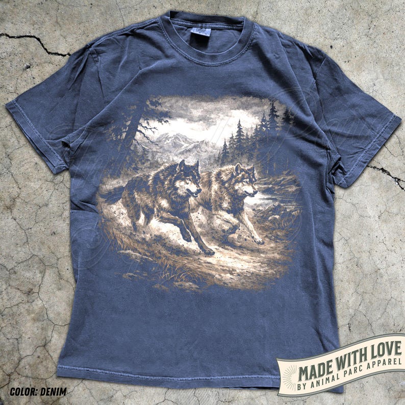 K&ouml;nnte beinhalten: Denimblaues T-Shirt mit einer Grafik von zwei W&ouml;lfen, die durch einen Wald laufen. Das Design umfasst B&auml;ume, Berge und einen bew&ouml;lkten Himmel. Das T-Shirt hat einen Rundhalsausschnitt und kurze &Auml;rmel. Der Text "MADE WITH LOVE BY ANIMAL PARC APPAREL" ist auf einem Banner aufgedruckt.