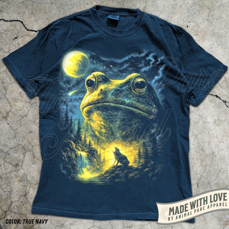 May include: A true navy short-sleeve t-shirt featuring a graphic print of a large frog head above a forest landscape with a waterfall, a glowing yellow moon, and a silhouette of a howling wolf against a starry night sky background.