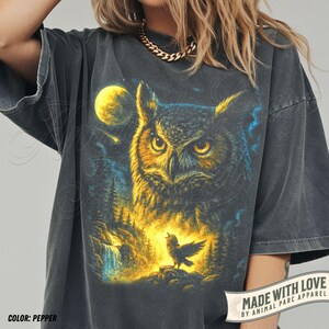May include: Pepper-coloured t-shirt with an owl illustration, full moon, and a small bird. The design uses yellow, blue, and black tones. The shirt has short sleeves and a relaxed fit. The text "MADE WITH LOVE" is visible on the bottom right.