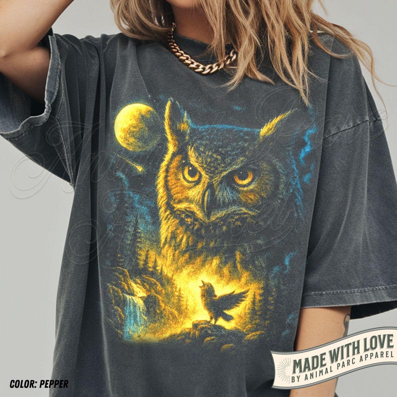May include: Pepper-coloured t-shirt with an owl illustration, full moon, and a small bird. The design uses yellow, blue, and black tones. The shirt has short sleeves and a relaxed fit. The text "MADE WITH LOVE" is visible on the bottom right.