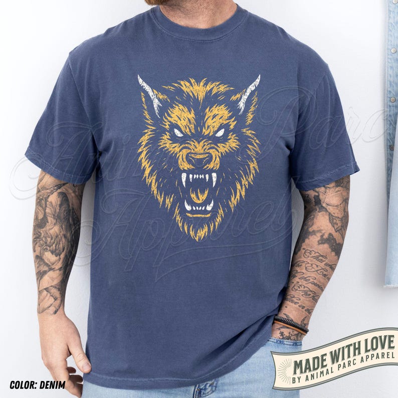 May include: Denim blue t-shirt with a yellow and white wolf head design. The wolf's snarling face is the central image. The text "MADE WITH LOVE BY ANIMAL PARC APPAREL" is printed on a banner. The shirt is a casual wear item.