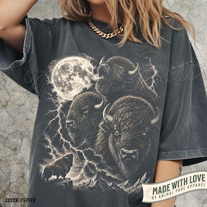 May include: A charcoal gray short-sleeve t-shirt featuring a graphic print of three bison heads, a full moon, and lightning bolts. A smaller bison stands on a rocky cliff in the foreground. The shirt has a distressed, vintage-style wash finish.