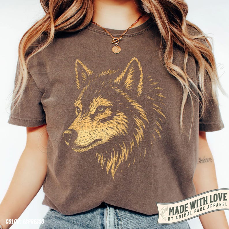 May include: A brown t-shirt featuring a detailed illustration of a wolf's head in shades of gold and brown. The shirt has a vintage, worn look. The text "MADE WITH LOVE BY ANIMAL PARC APPAREL" is printed on a banner.